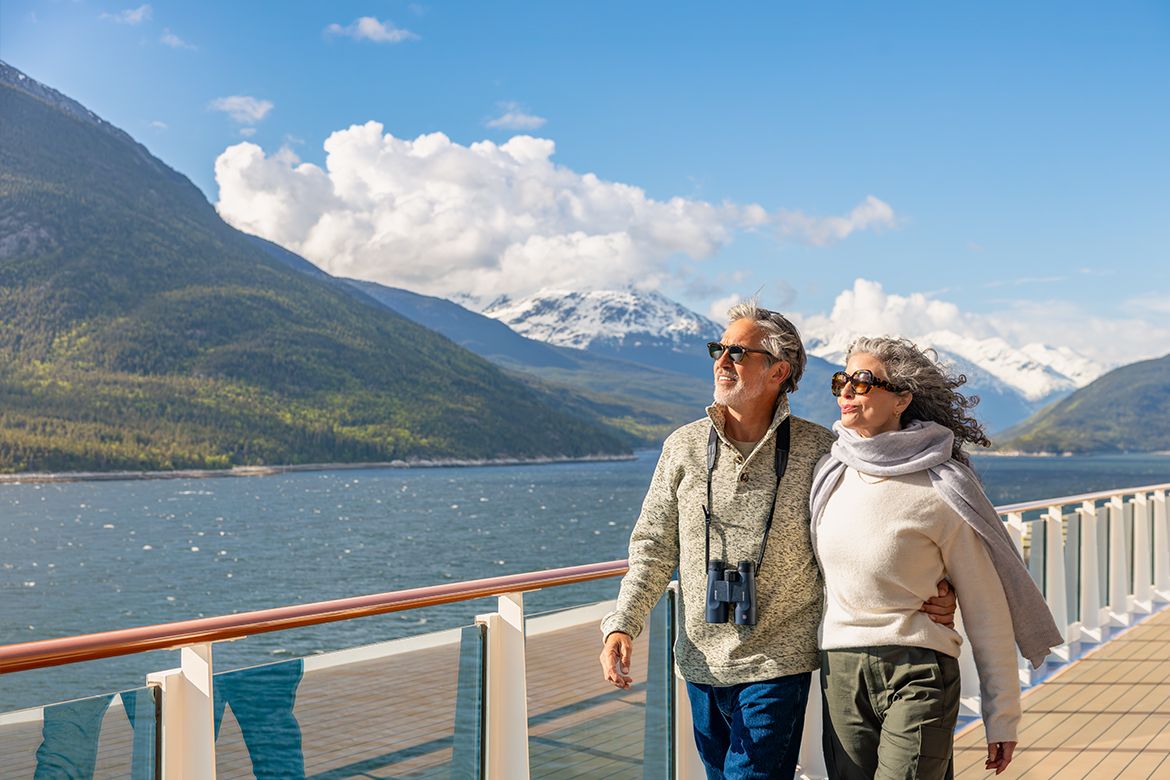 Couple walking on a cruise ship deck beside a mountain-lined waterway.