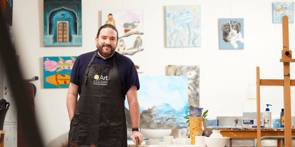 Gordon standing inside QArt Studio surrounded by original artworks, canvases and creative workspaces