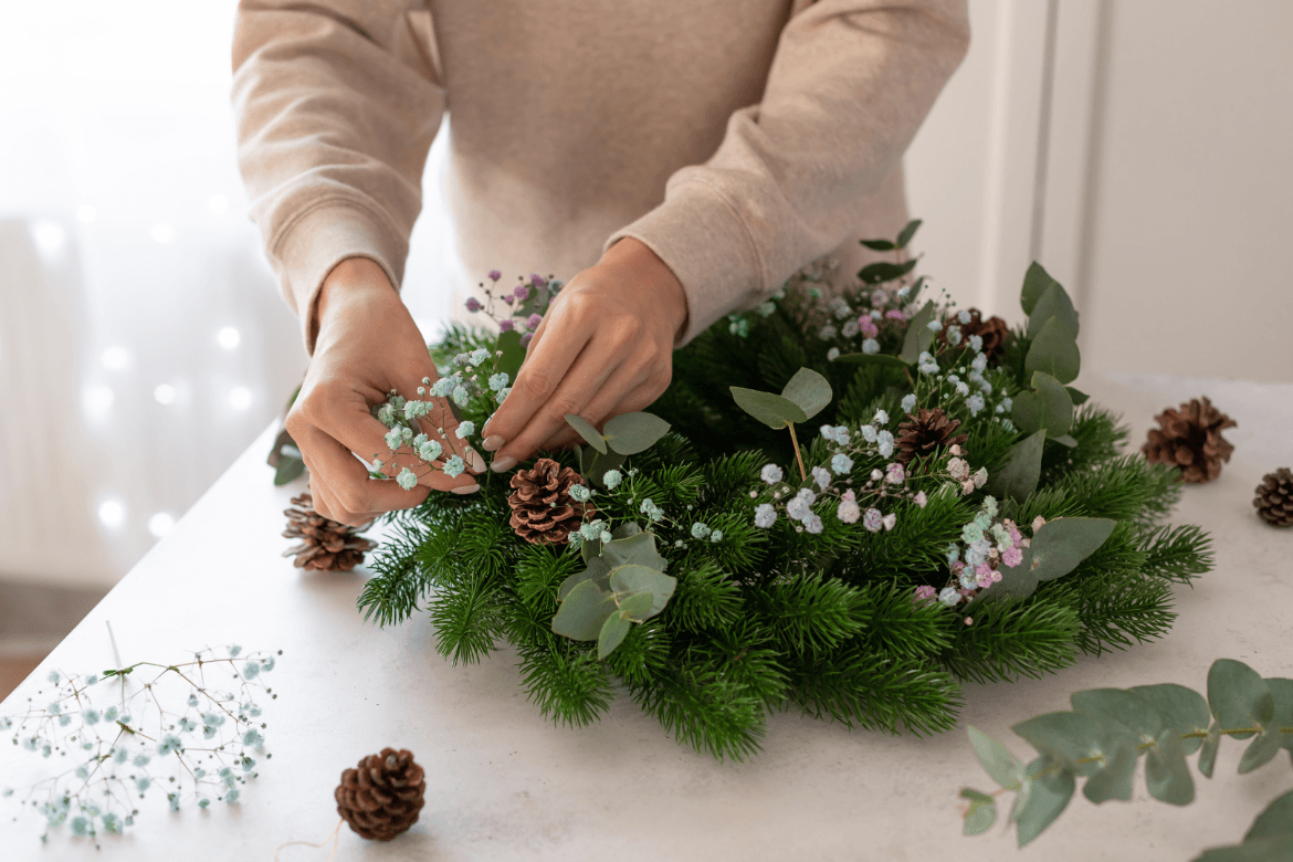 Hands decorating a wreath with baby's breath flowers, pinecones, and eucalyptus on a white table