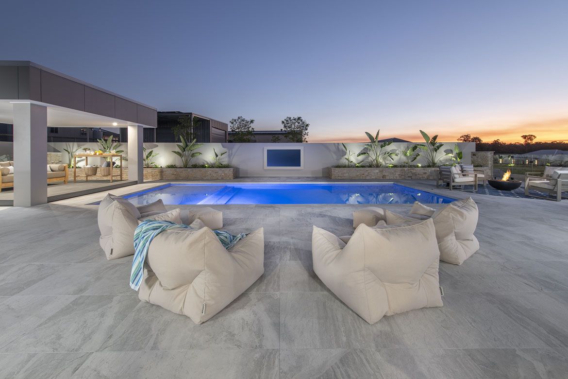 Outdoor pool area at dusk with glowing blue water, beige bean bag chairs, a fire pit, and a covered lounge area surrounded by plants.