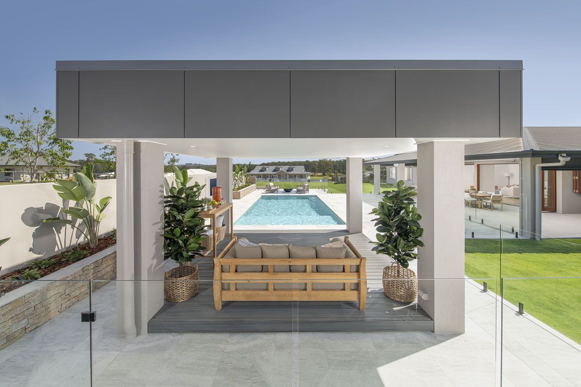 Covered outdoor seating area with a wooden couch overlooks a long swimming pool. Surrounded by planters and glass fencing, leading to a spacious lawn and a modern house.