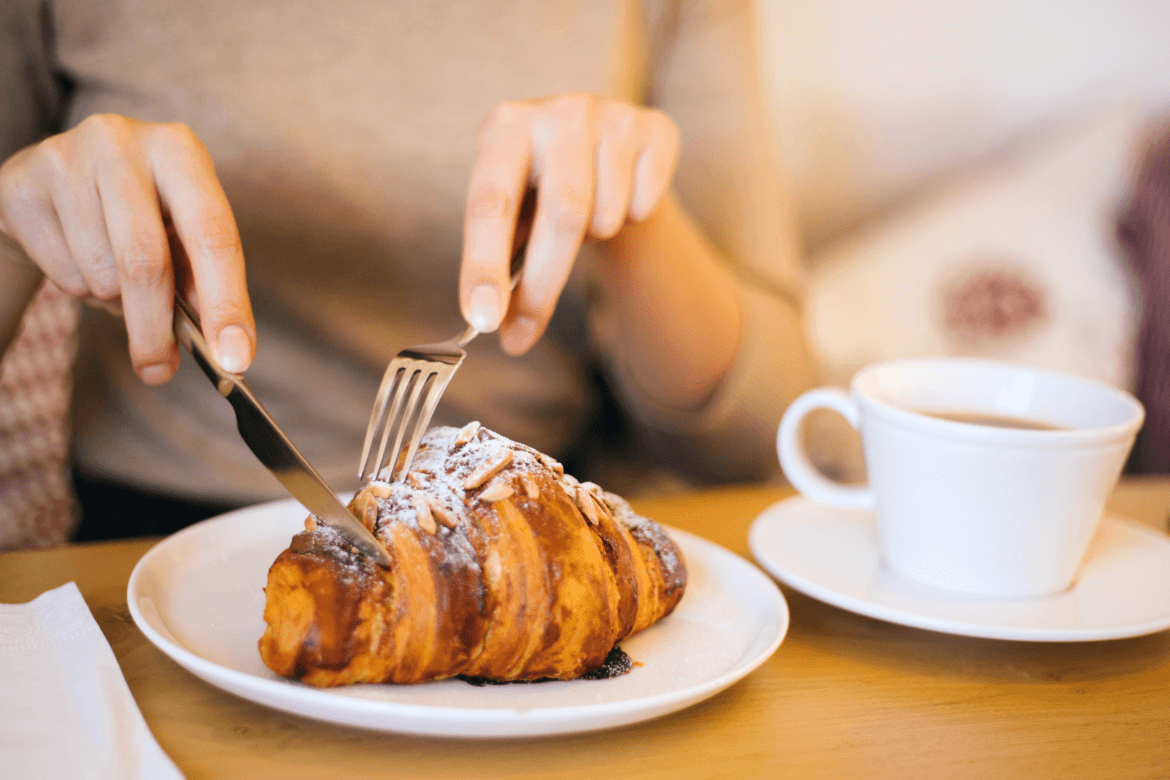 A woman enjoys a flaky croissant, holding a fork in one hand and a knife in the other, ready to take a bite.