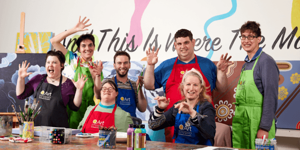 Group of QArt Studio artists smiling and posing together in their Melbourne studio, surrounded by colourful artworks and painting materials