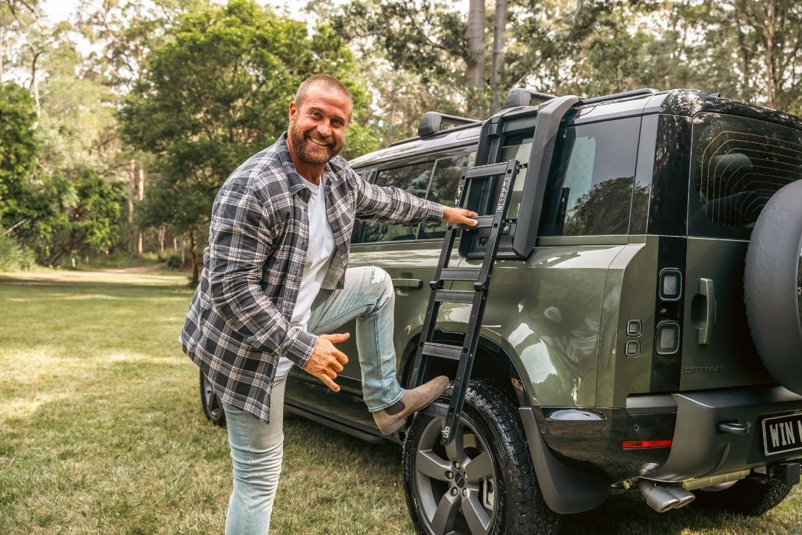 Person using deployable roof ladder on a green V8 Land Rover Defender in a bushland setting